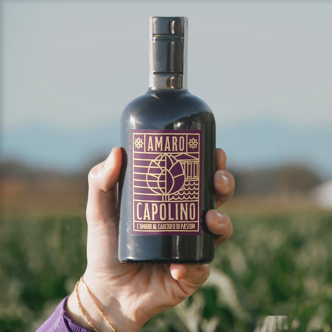 una mano che impugna la bottiglia di amaro capolino con sfondo di un campo sfocato