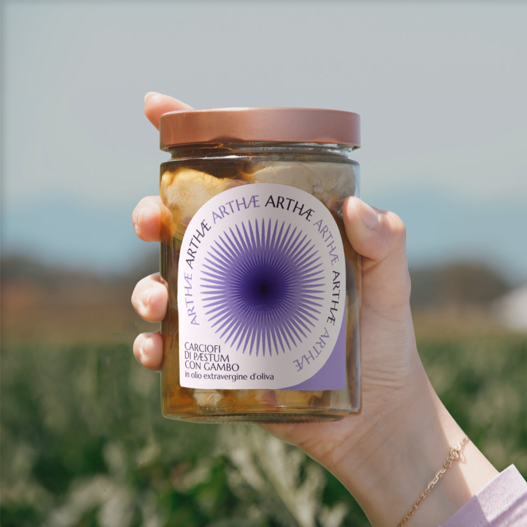 una mano che impugna il barattolo di sott'oli linea Arthae con sfondo di un campo sfocato